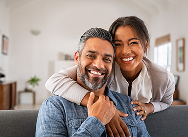 Portrait of multiethnic couple embracing and looking at camera sitting on sofa. Smiling african american woman hugging mid adult man sitting on couch from behind at home. Happy mature mixed race couple laughing at home.
